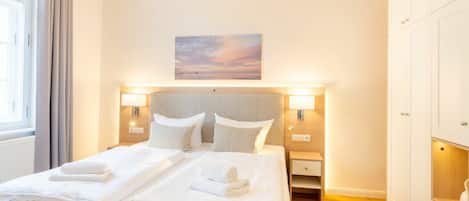A hotel room with a bed, two bedside tables, a nightstand, a wall-mounted lamp, a painting, and a wardrobe.