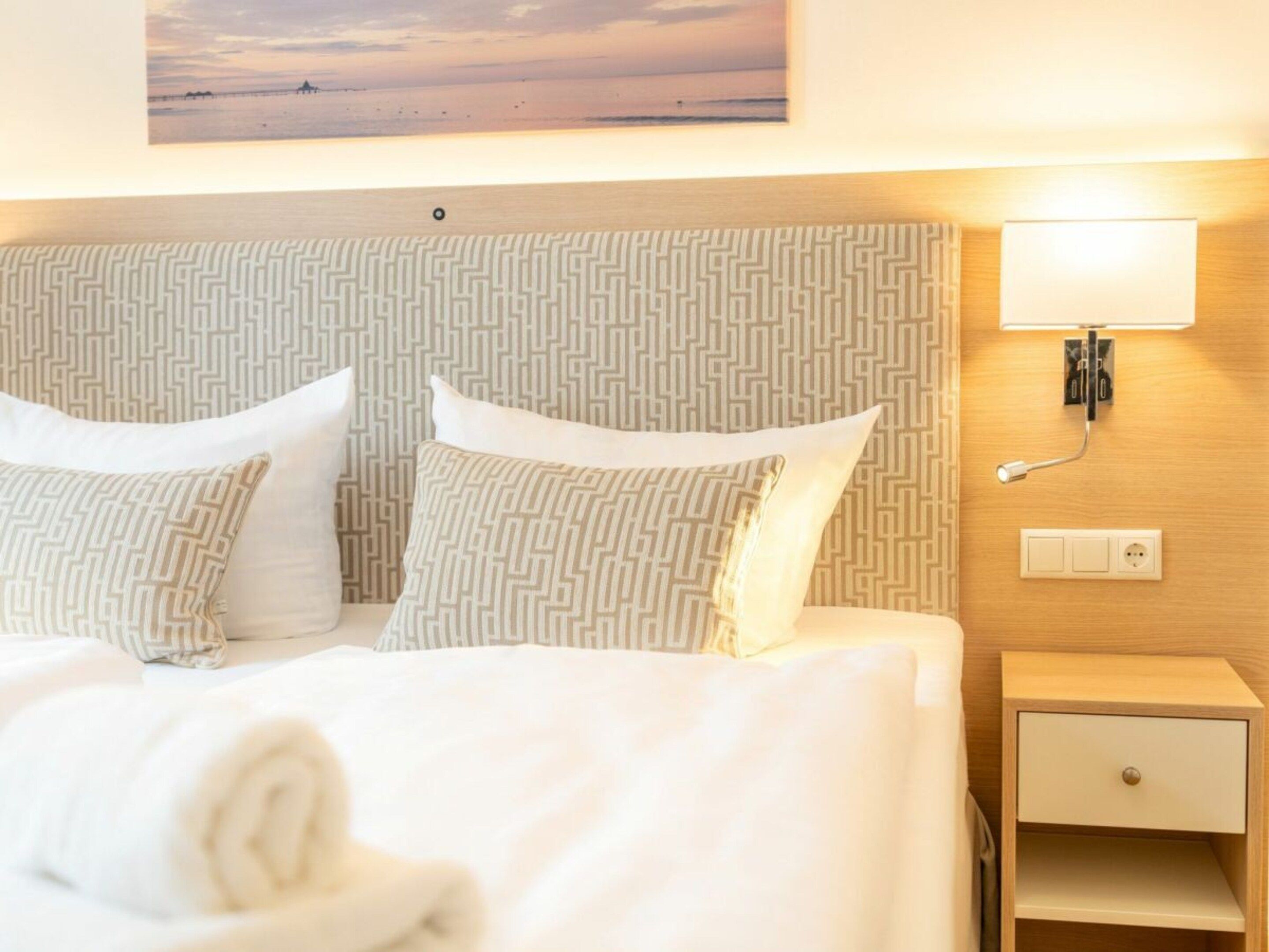 Une chambre d’hôtel comprenant un lit, des oreillers, une table de chevet, une lampe et une prise électrique fixée au mur.