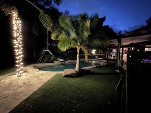 Ein Außenbereich bei Nacht mit einem Schwimmbecken, Palmen und einer Lichterkette.