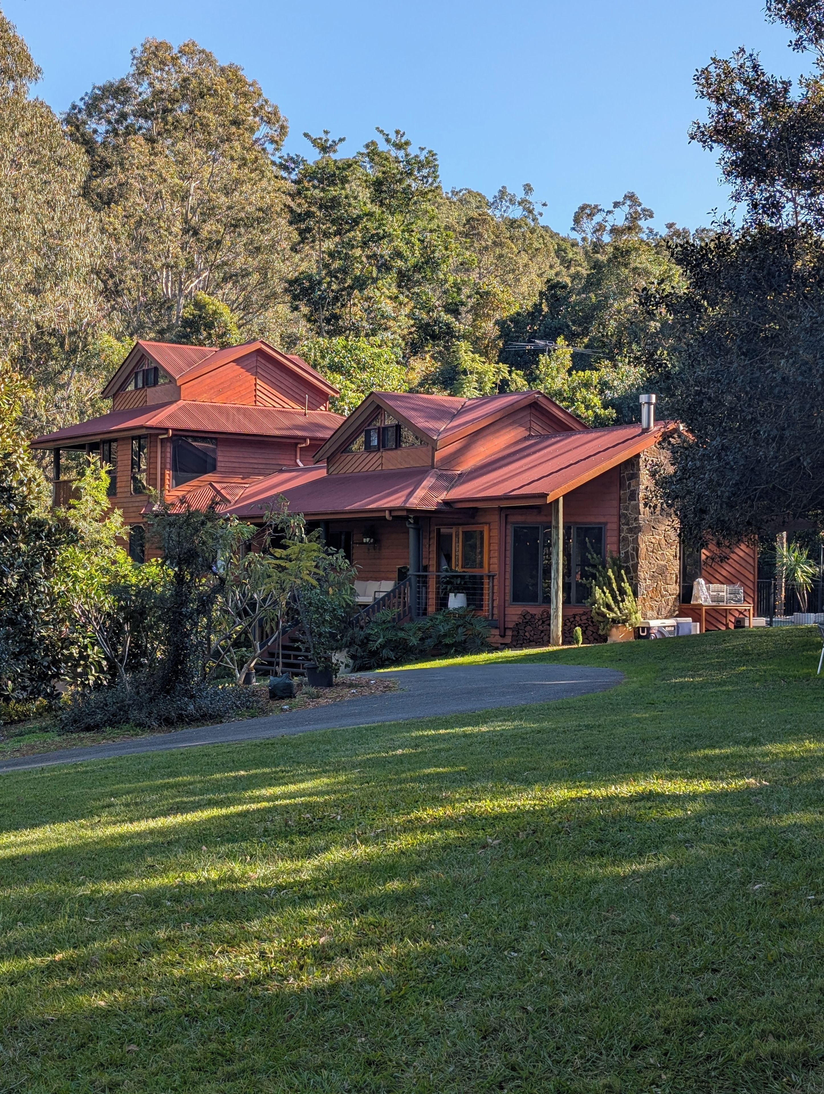 Une maison à deux étages, avec un toit rouge, entourée d’une végétation luxuriante et d’arbres.