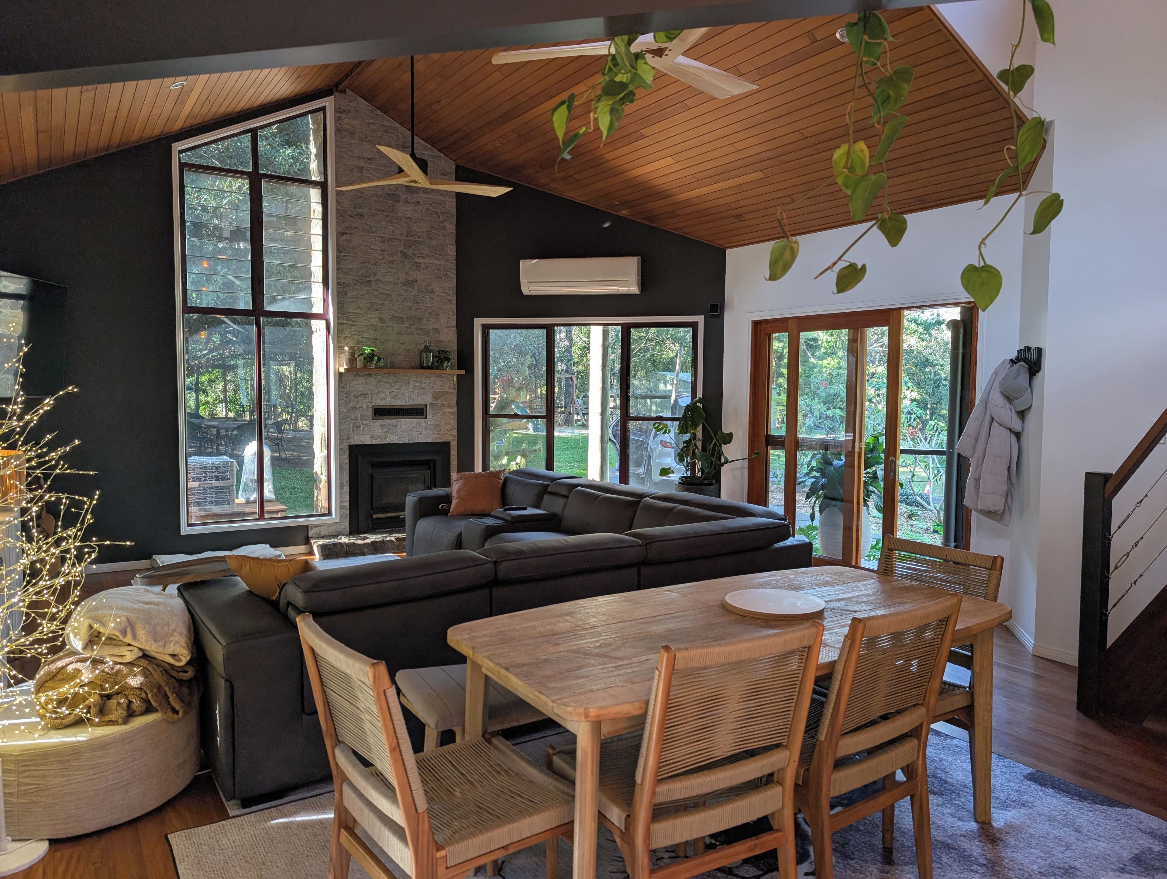 A modern living room with a sectional sofa, dining table, and chairs. There is a fireplace, large windows, and a ceiling fan.