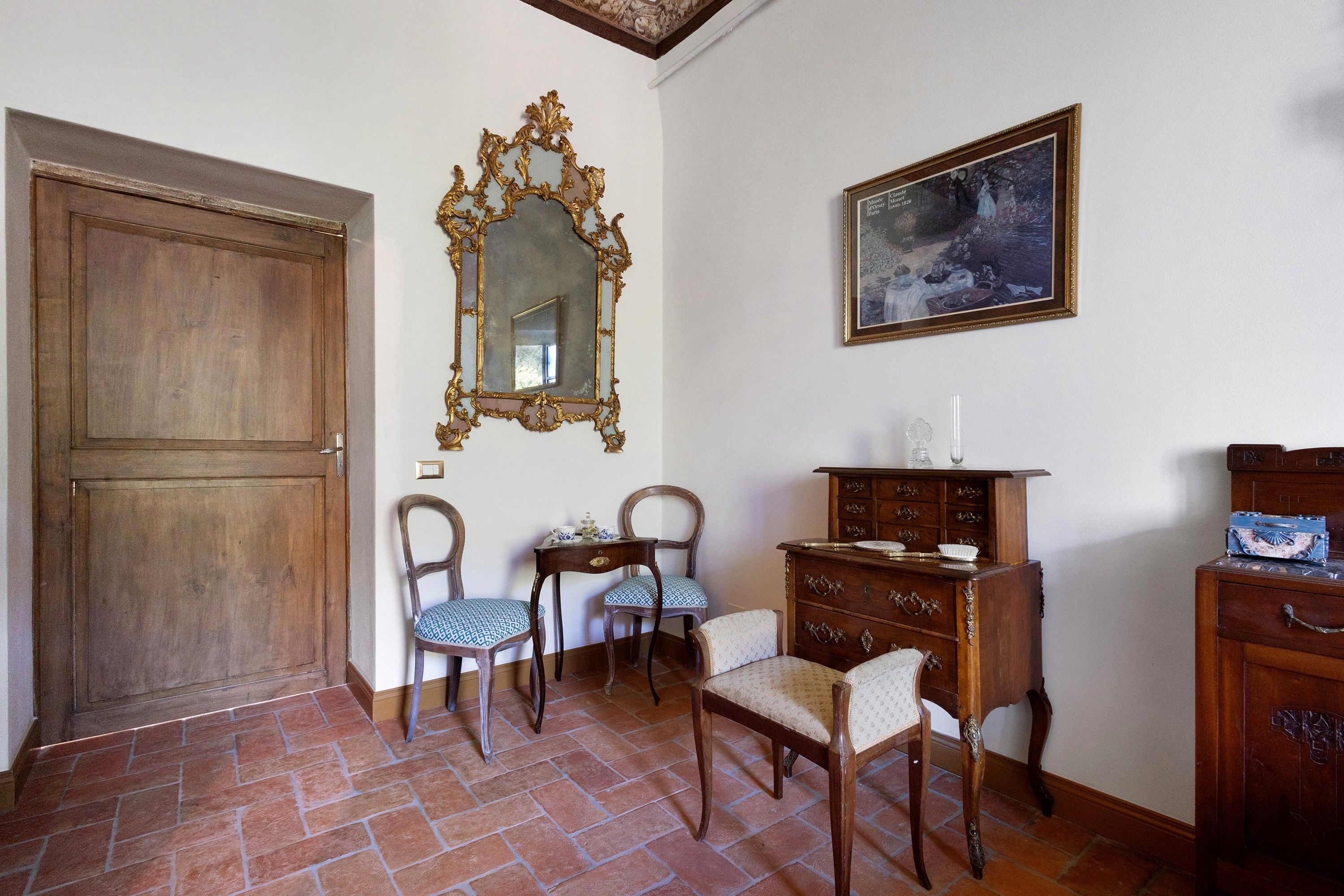 A room with a wooden door, two chairs with patterned upholstery, a small table with a decorative item, a wooden dresser with drawers, and a framed painting on the wall.