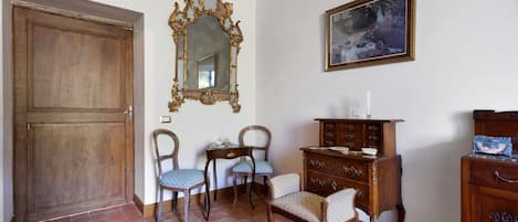 Una habitación con puerta de madera, dos sillas con tapicería estampada, una mesita con un objeto decorativo, un aparador de madera con cajones y un cuadro enmarcado en la pared.