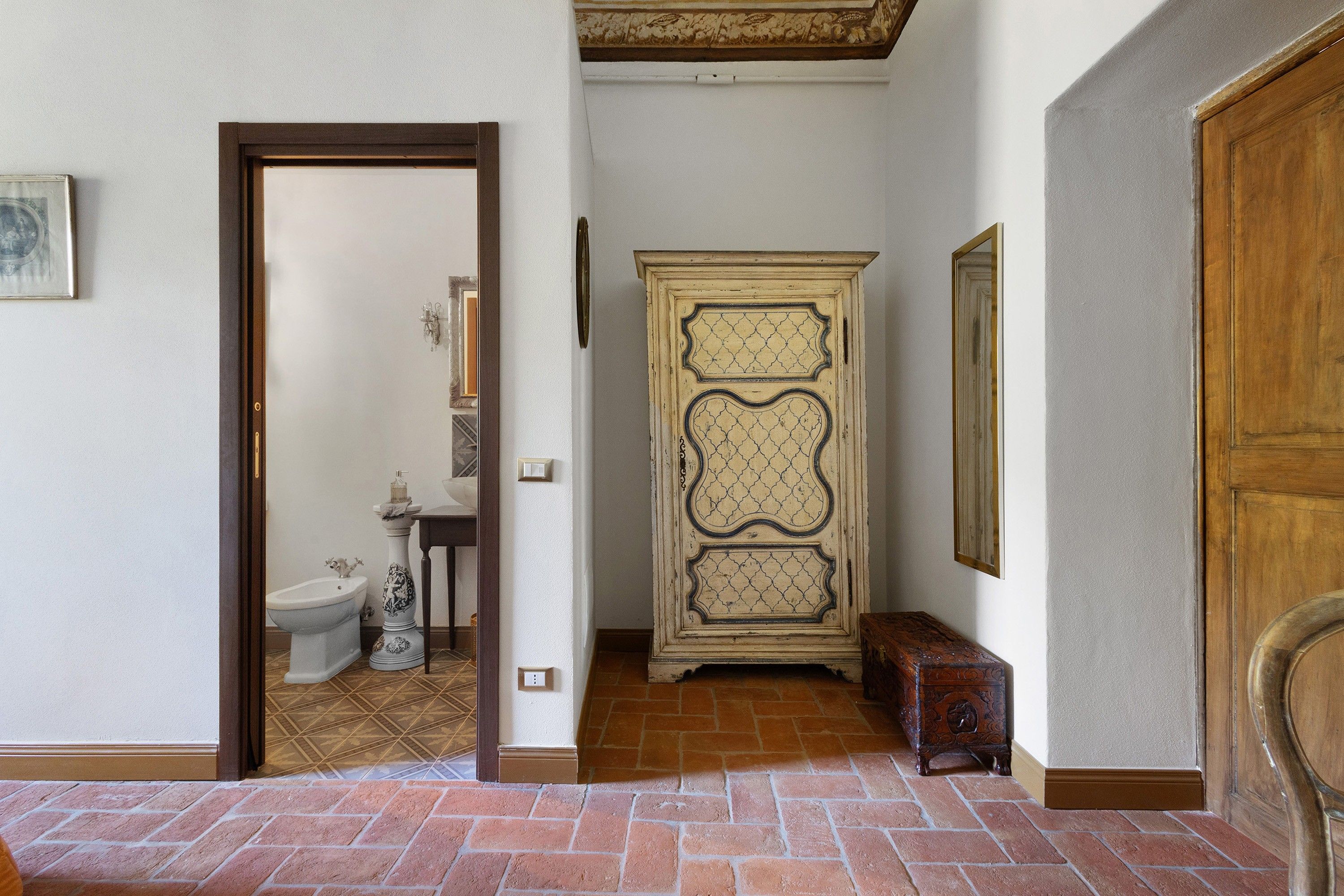 A hallway with a wooden door, a white wall, a tiled floor, a white toilet, and a cabinet with decorative patterns.