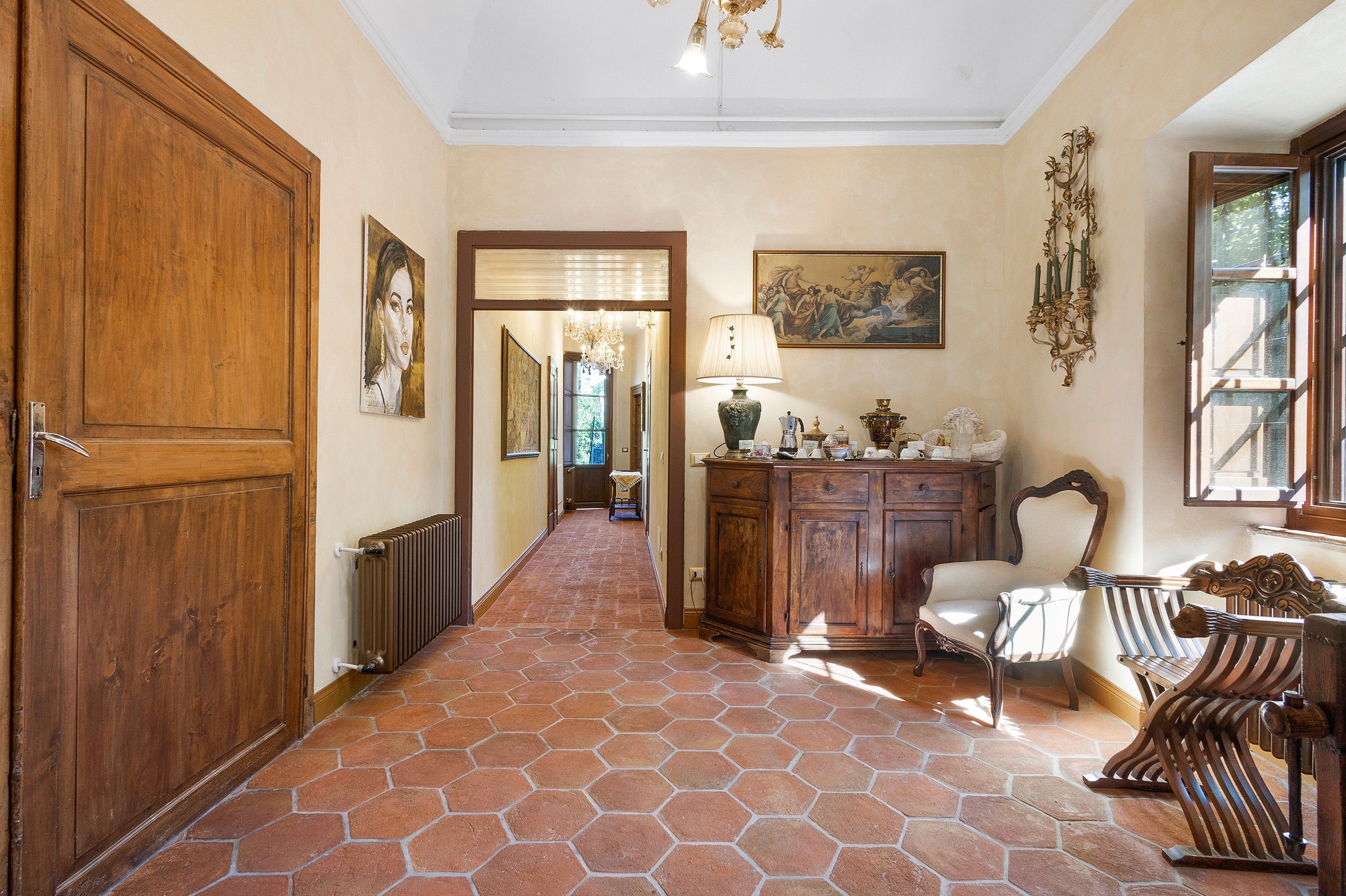Un couloir avec une porte en bois, un radiateur, une table avec une lampe, une chaise et un meuble contenant des objets décoratifs.