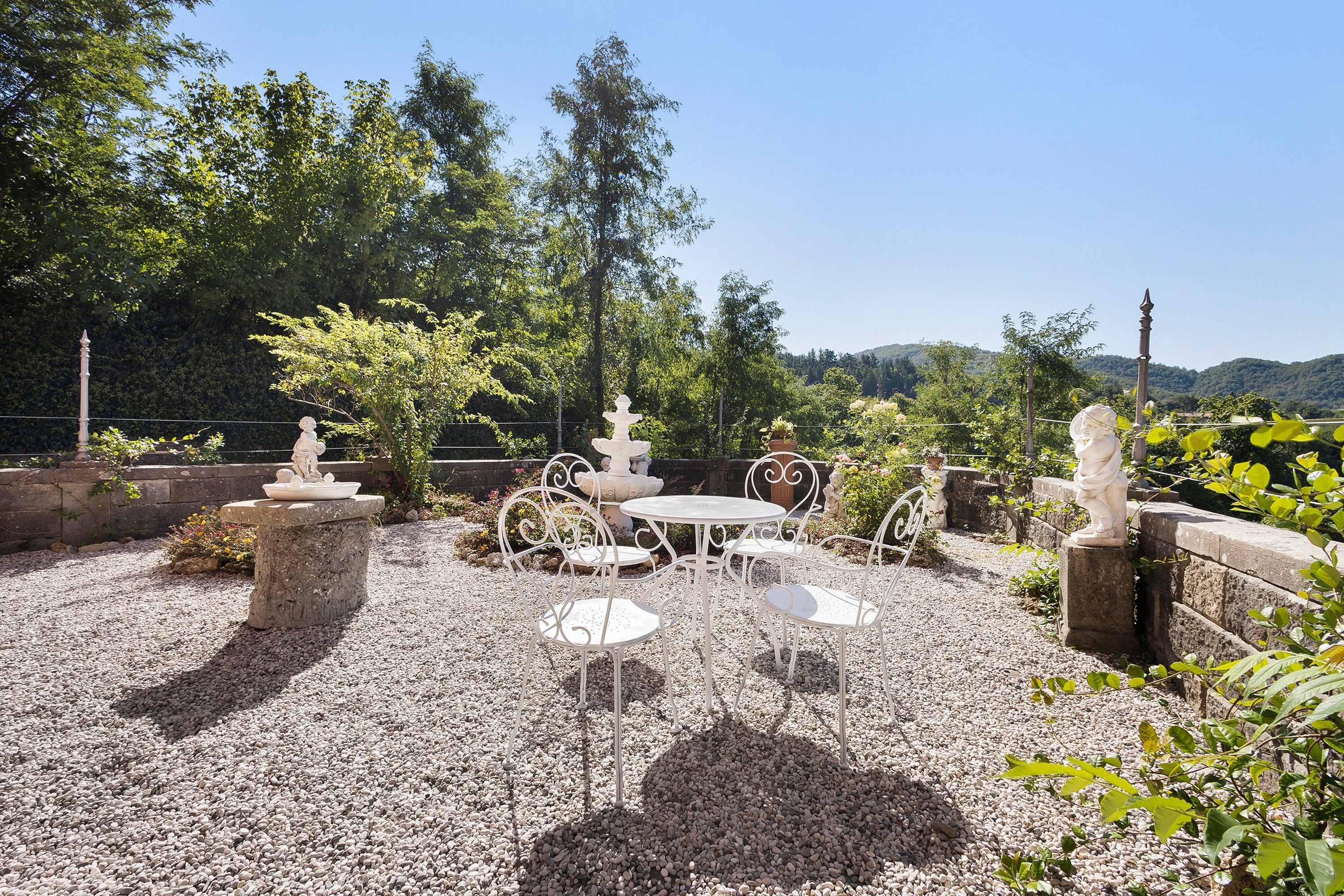 Eine Terrasse im Freien mit Tisch und Stühlen, umgeben von Steinmauern und viel Grün.