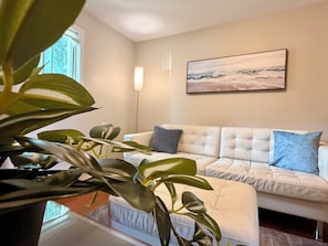 Un salon avec un canapé blanc, un coussin bleu, un lampadaire et un tableau représentant une scène de plage.