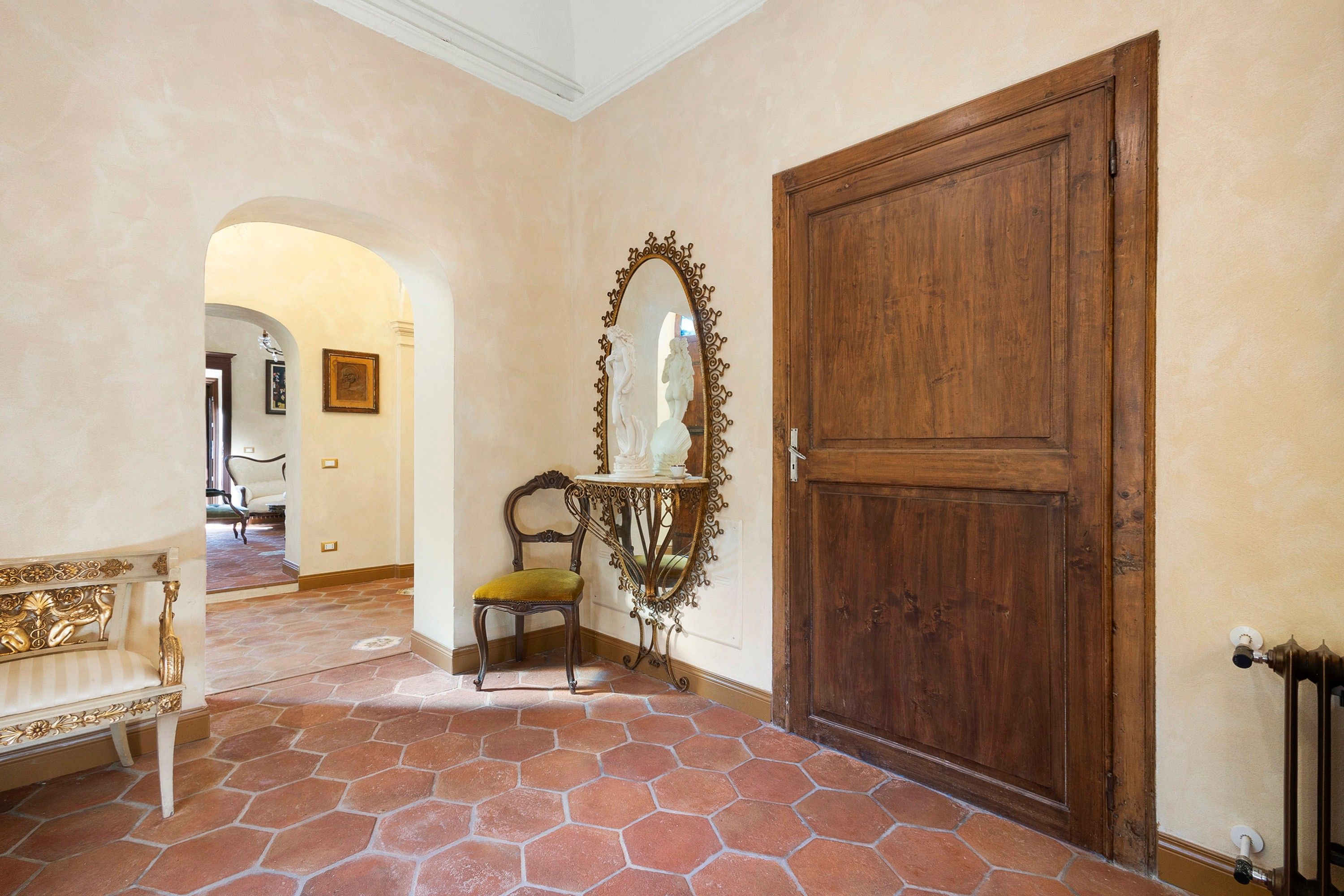 Un couloir avec une grande porte en bois, un miroir doté d’un cadre orné, une chaise avec un coussin jaune et un banc avec un dossier décoratif.