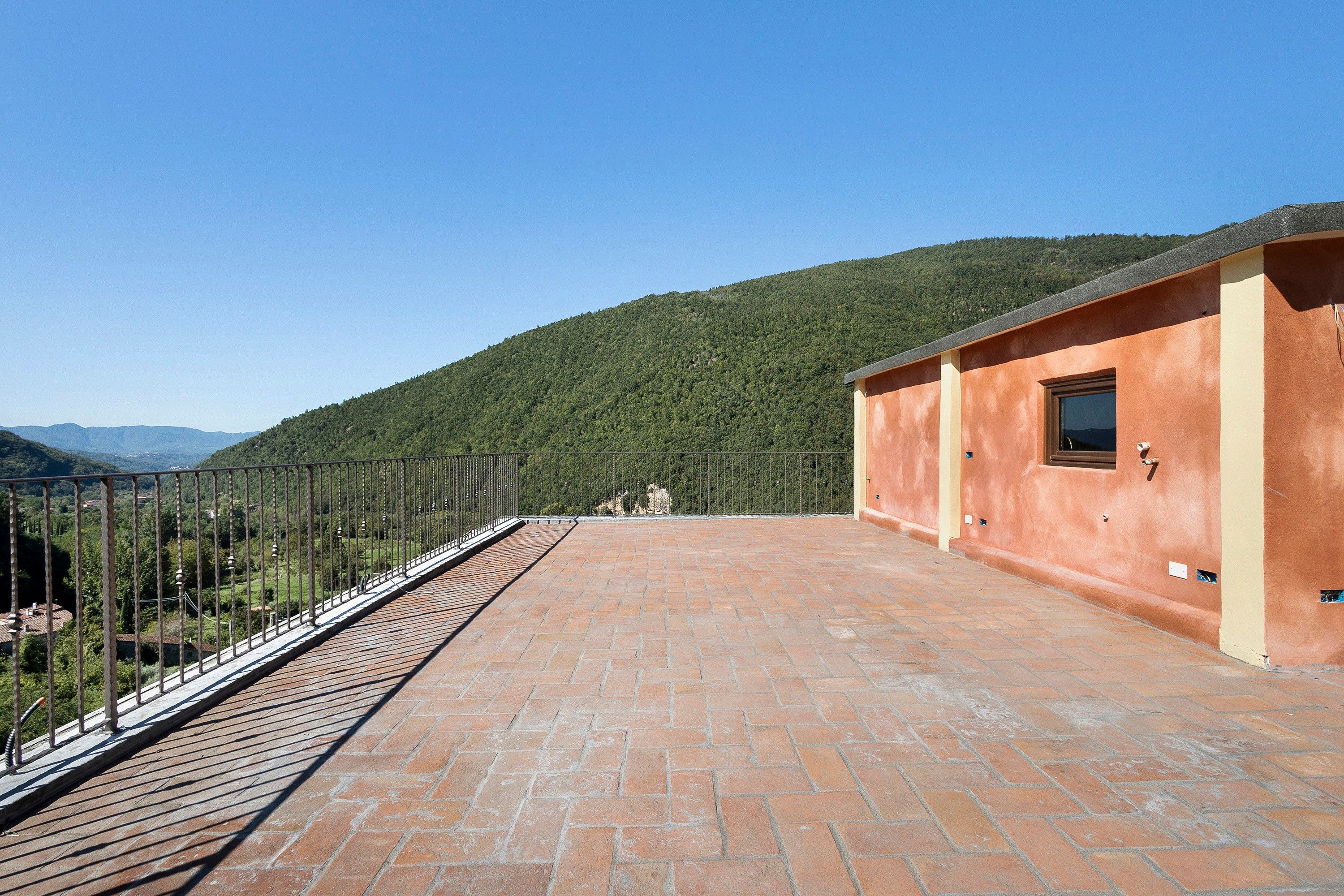 Een terras op het dak met een metselstenen vloer, een metalen hek en uitzicht op een groene berg.