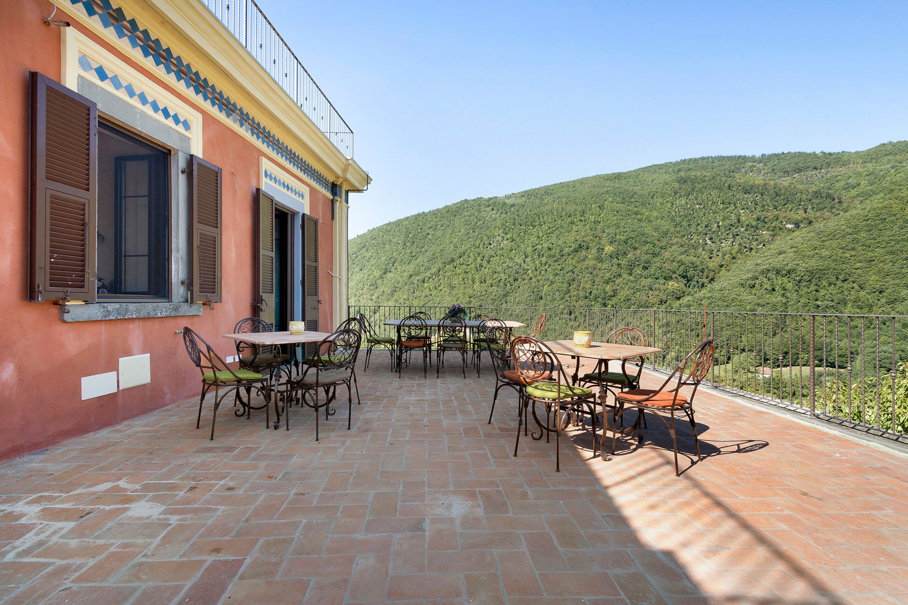 Un patio exterior con mesas y sillas, un edificio rojo con persianas y vistas a una ladera verde.