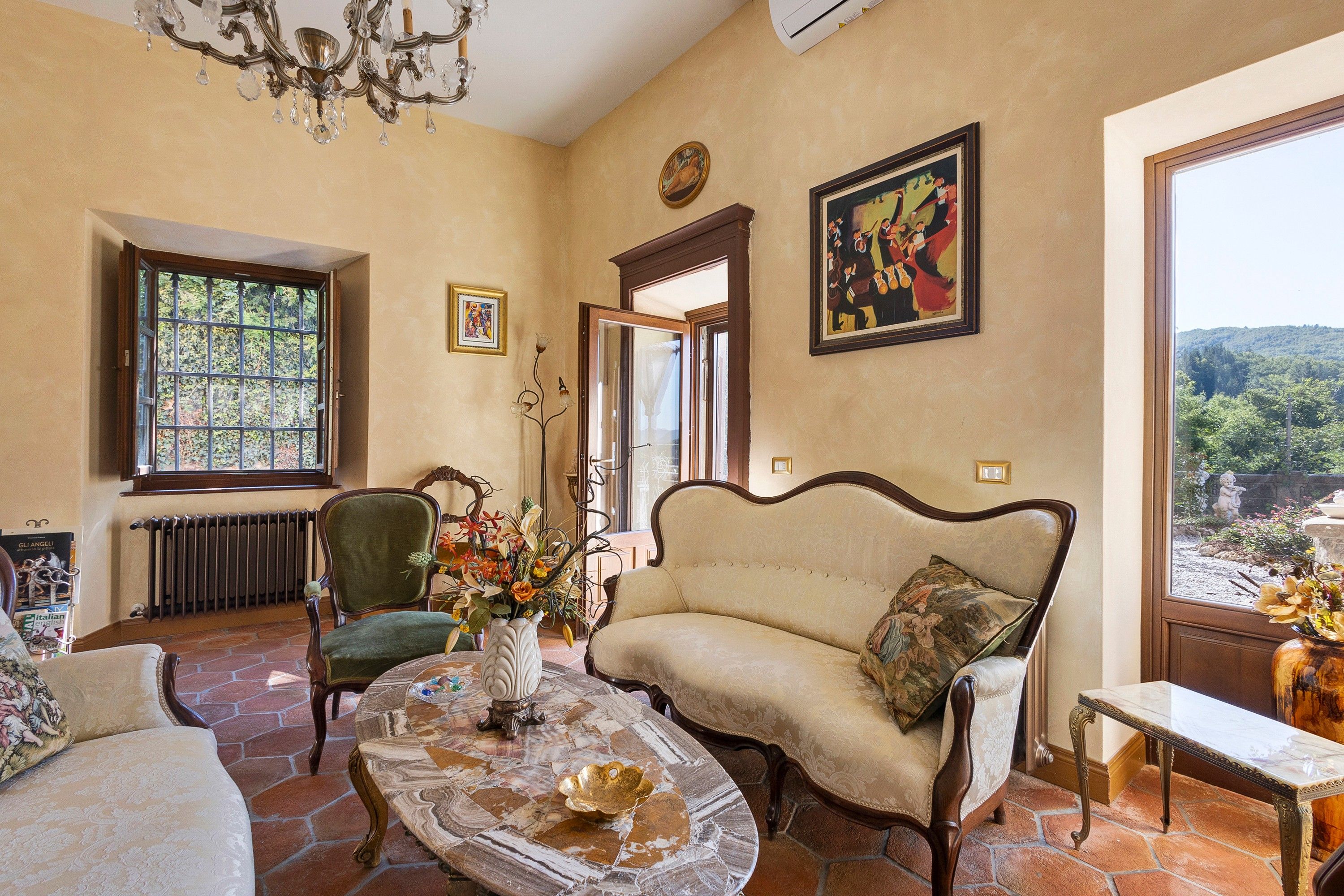 A spacious room with a large window, a green armchair, a beige sofa, a marble coffee table, and a chandelier.
