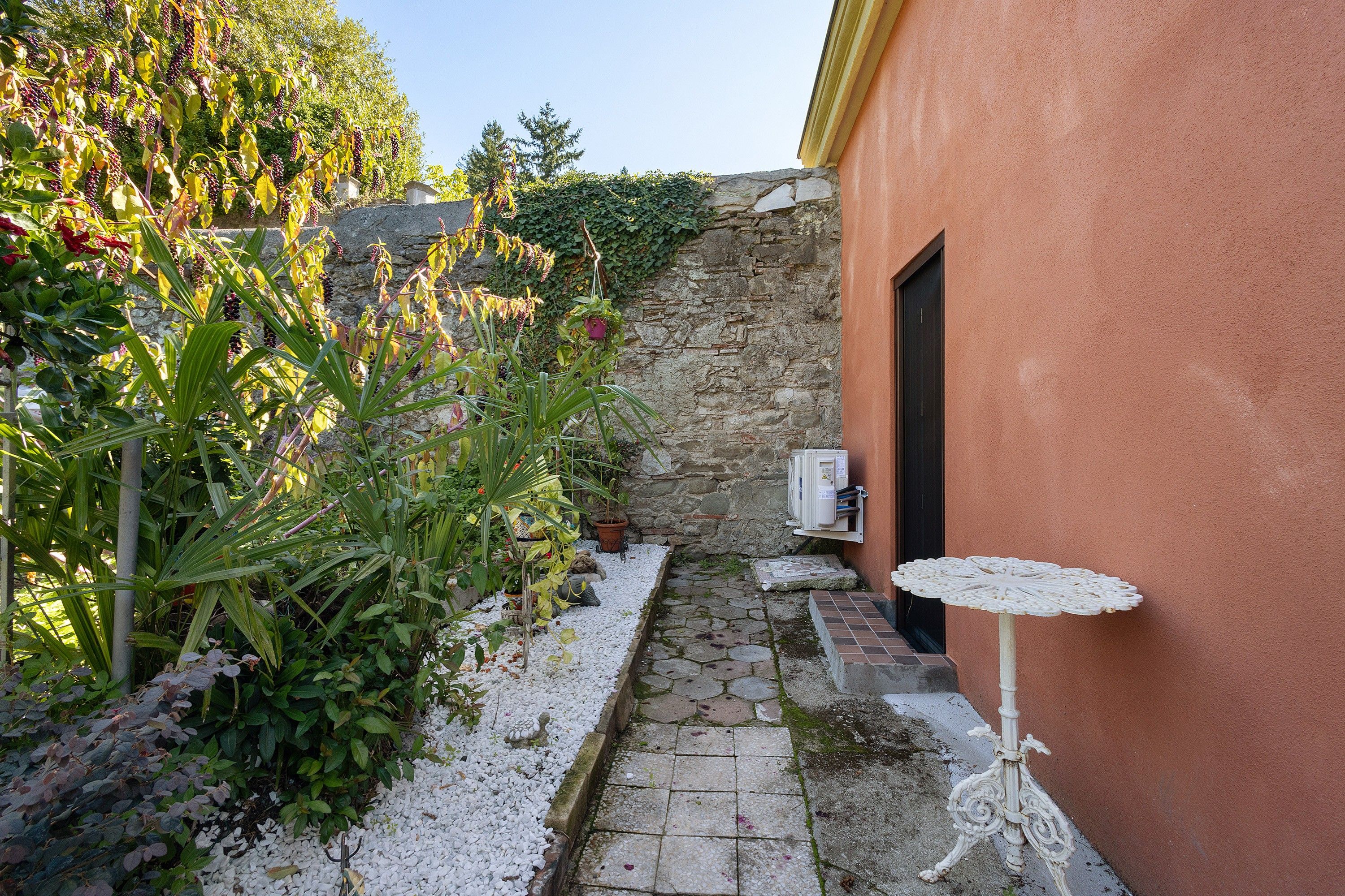 Un stretto sentiero in pietra conduce a una porta, circondato da una vegetazione lussureggiante e un piccolo tavolo bianco.