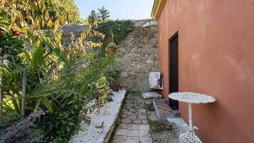 Un stretto sentiero in pietra conduce a una porta, circondato da una vegetazione lussureggiante e un piccolo tavolo bianco.