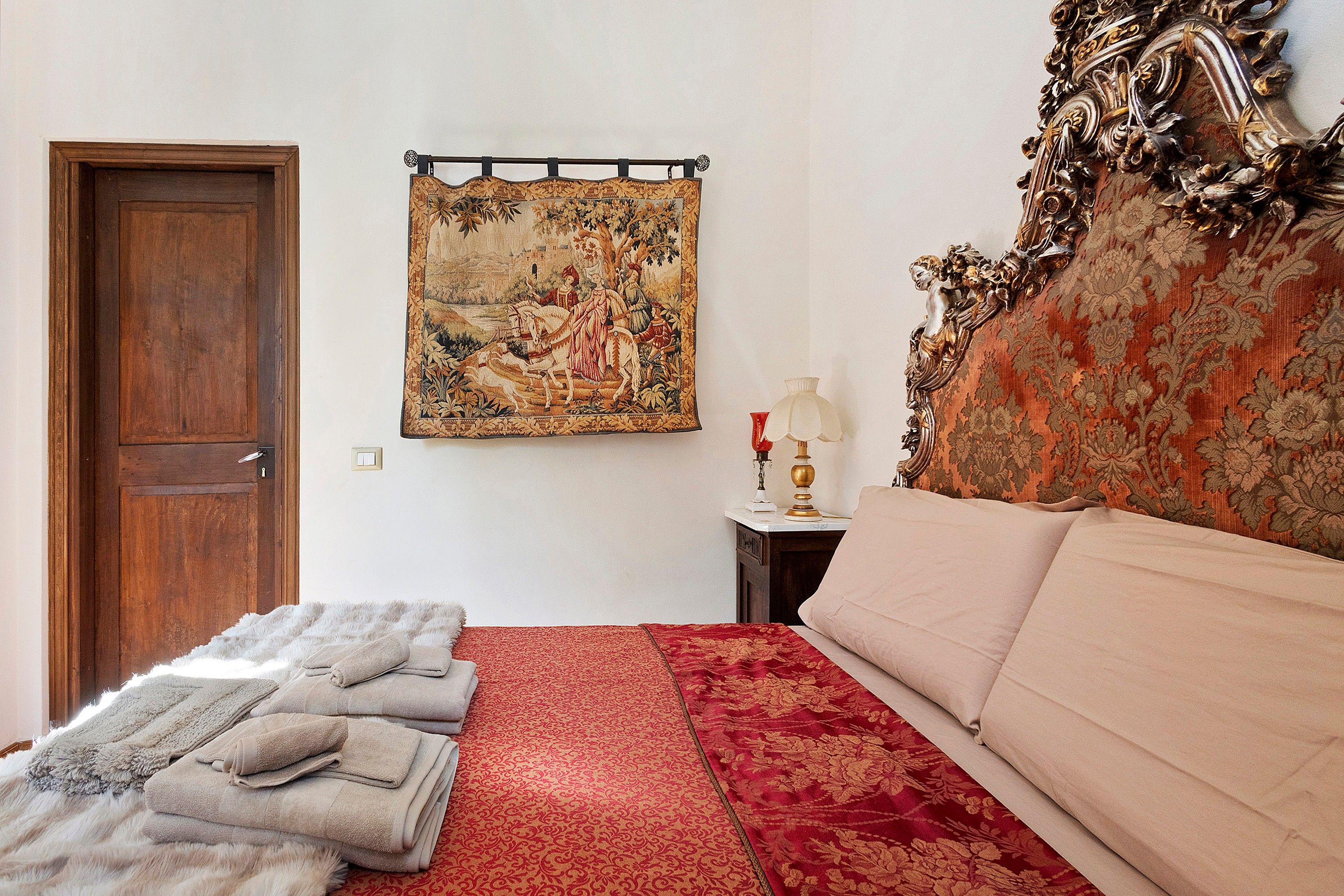 A bedroom with a wooden door, a tapestry, a bed with a red and beige bedspread, a nightstand with a lamp, and a decorative headboard.