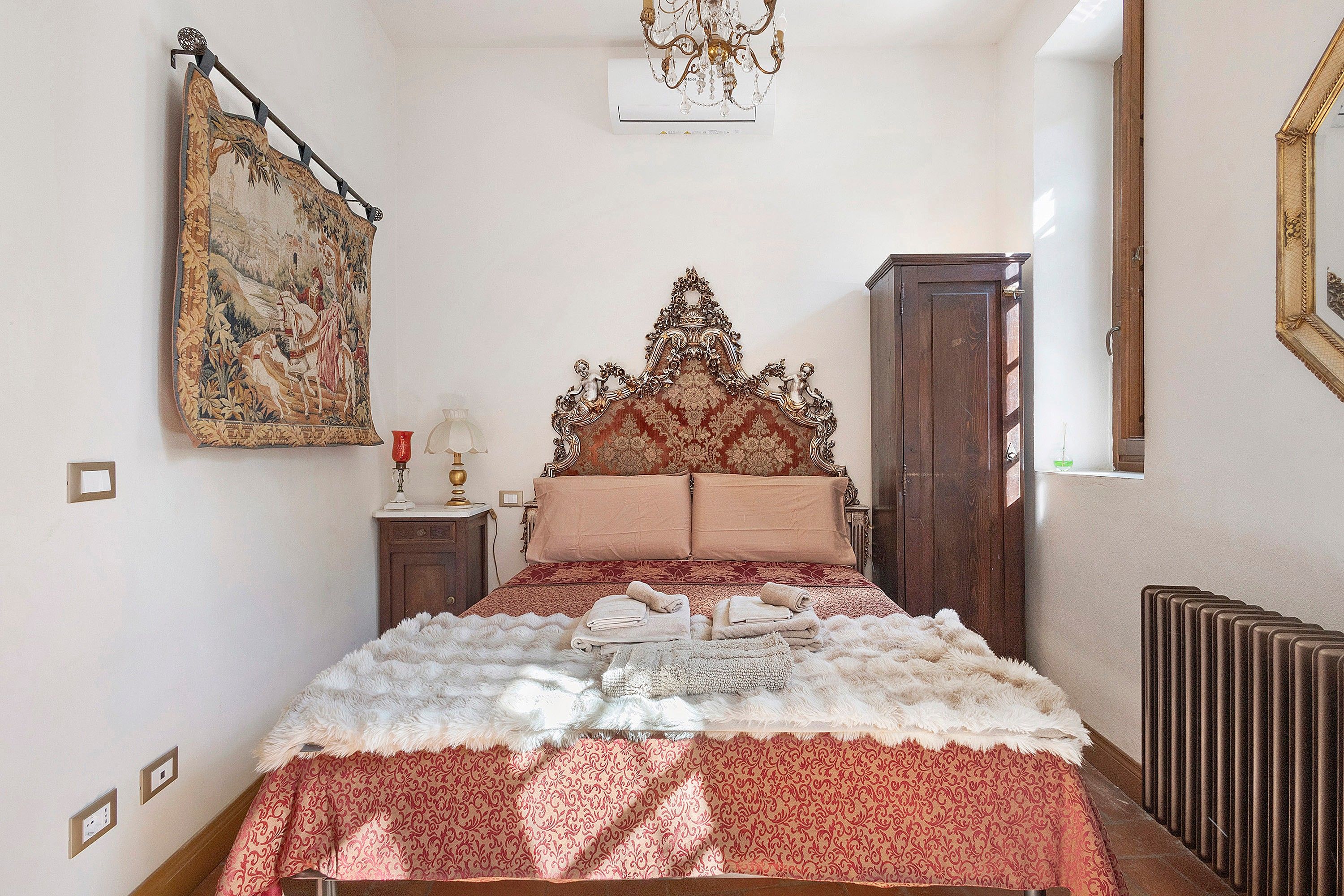 Une chambre avec un grand lit, une armoire en bois, une tapisserie au mur et un lustre.