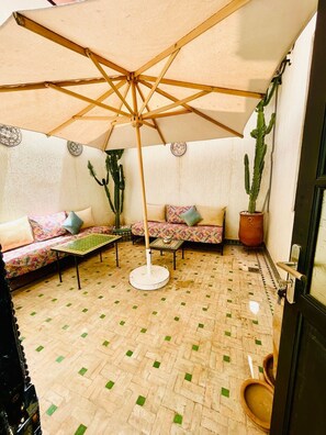 A cozy patio area with a wooden floor, a cushioned sofa, a round table, and a large umbrella providing shade. - Riad Douirtna (Marrakech)
