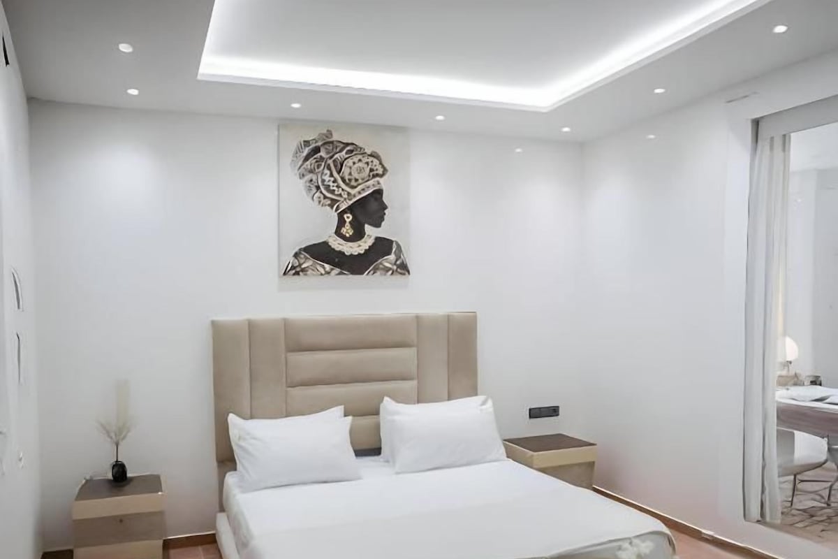 A modern bedroom with a large bed, bedside tables, a bench, and a wall-mounted artwork.