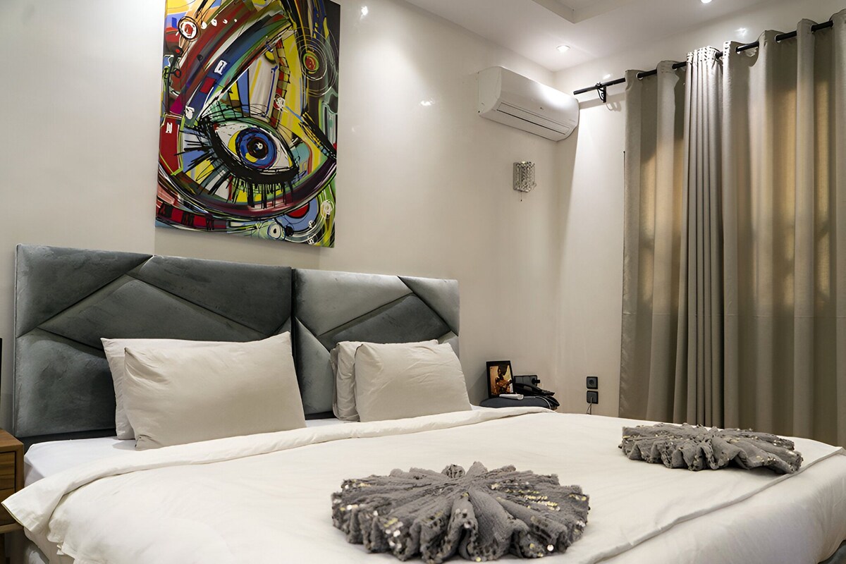 Un dormitorio moderno con una cama grande, un cojín decorativo, un aire acondicionado de pared y un cuadro.