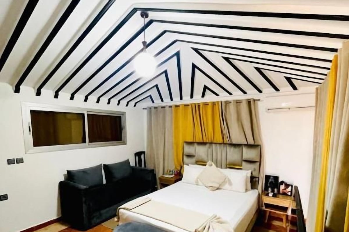 A bedroom with a bed, a bench, a window, and a ceiling with geometric patterns.