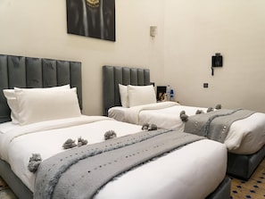 A hotel room with two beds, each with a grey headboard, white bedding, and decorative pillows. There is a wall-mounted phone and a small table with a bottle and a glass. - Riad Douirtna (Marrakesh-Safi)