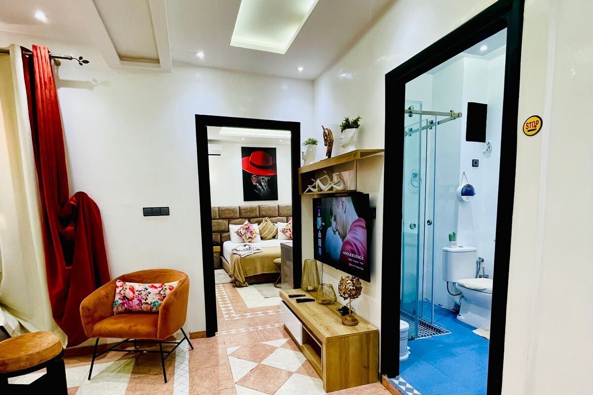 Una habitación de hotel moderna con piso a cuadros, un sofá, un televisor y un baño visible a través de una puerta de cristal.