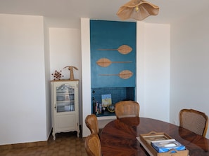 Uma área de refeições com uma mesa de madeira redonda, duas cadeiras de vime, um armário com portas de vidro e uma parede azul com detalhes decorativos.