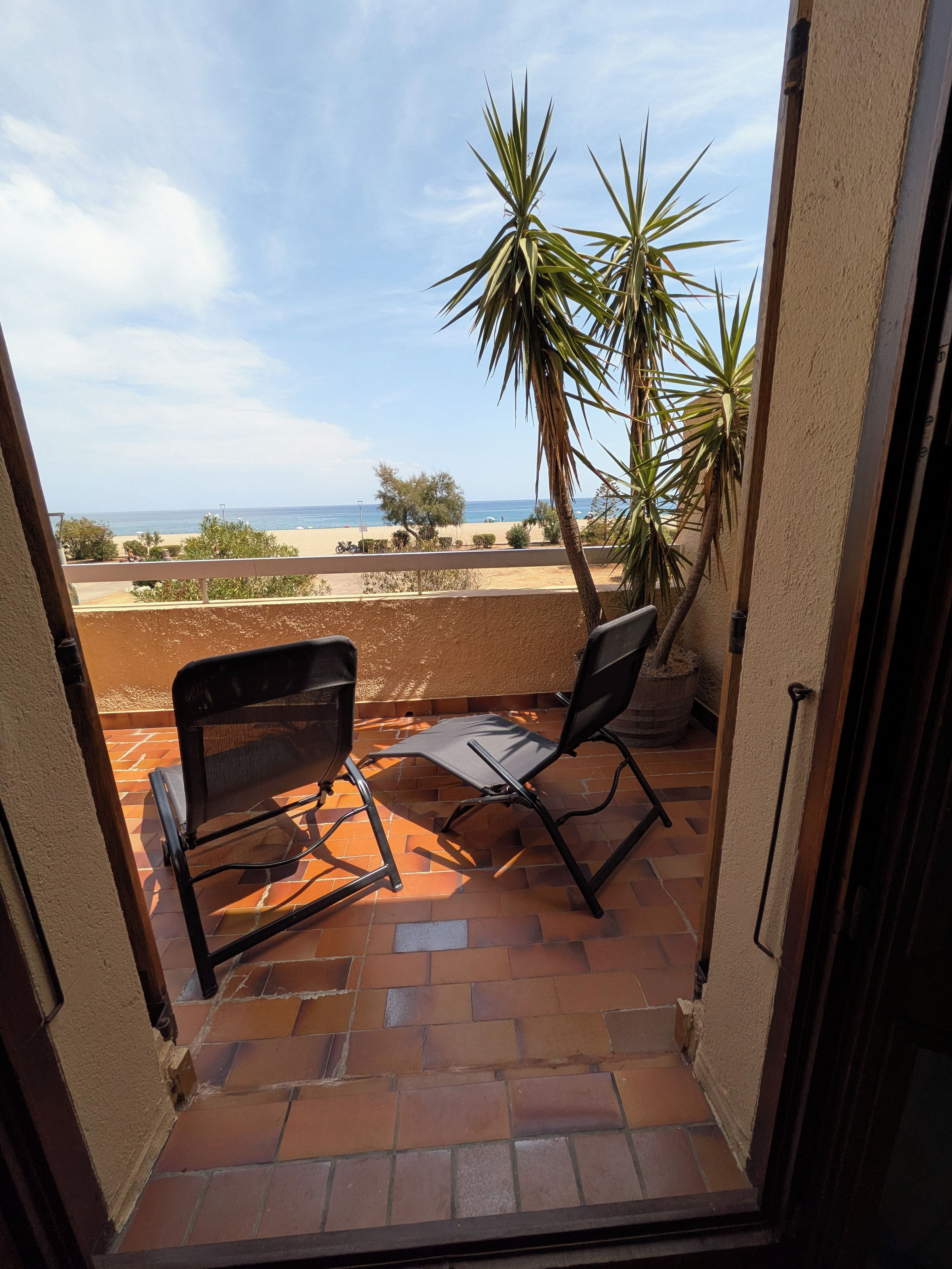 A balcony with two chairs and a table, overlooking a beach and palm trees.