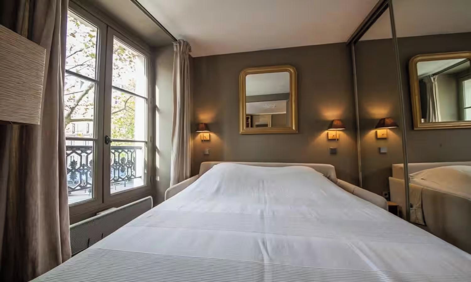 Een hotelkamer met een groot bed, twee wandlampen, een spiegel en een raam met gordijnen.