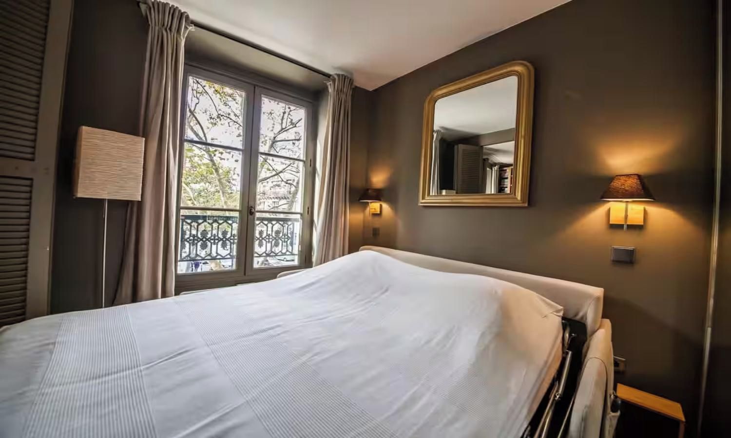 A hotel room with a bed, a window with curtains, a wall-mounted mirror, and two wall sconces.