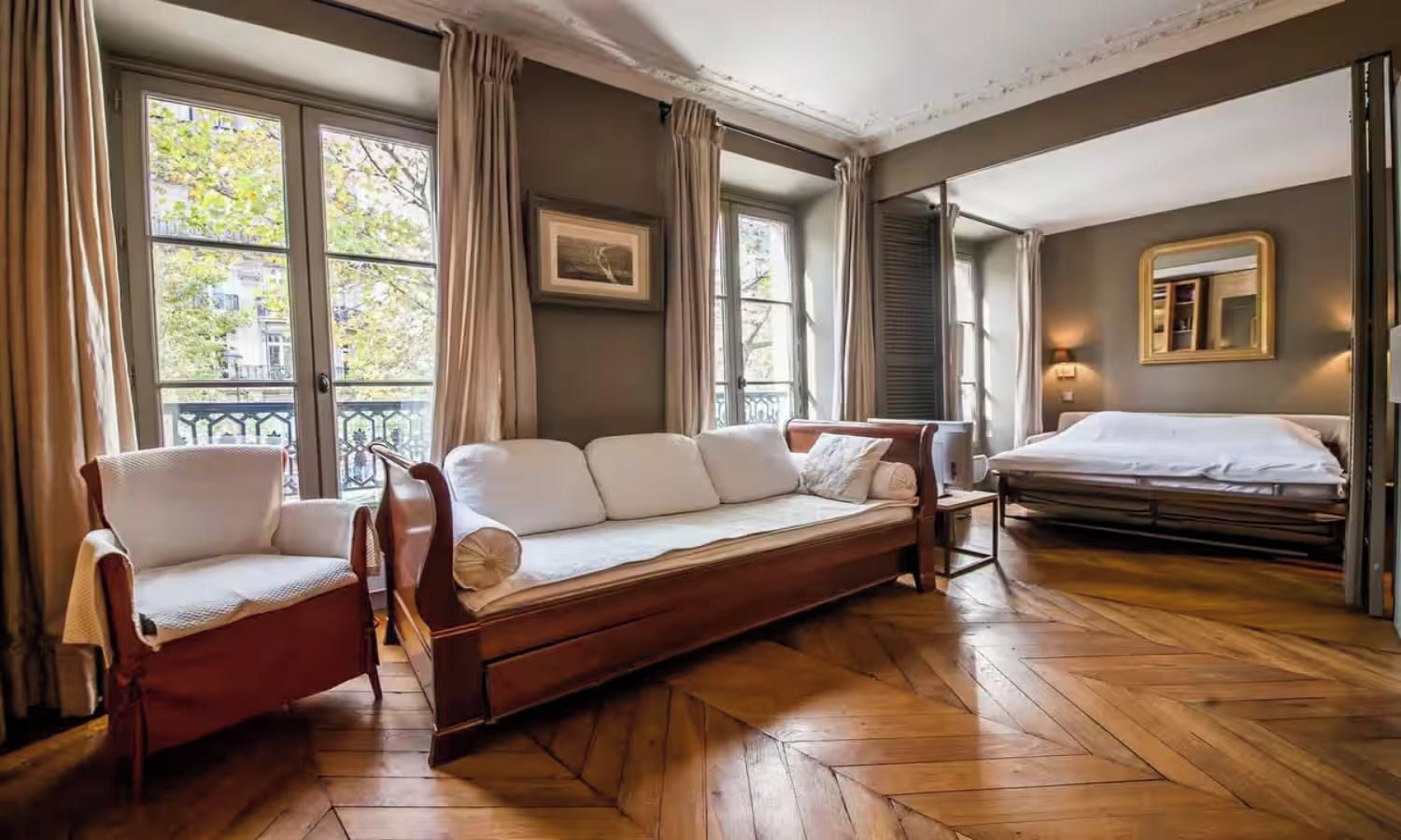 Una habitación con piso de madera, un sofá, una silla, una cama y ventanas grandes con cortinas.