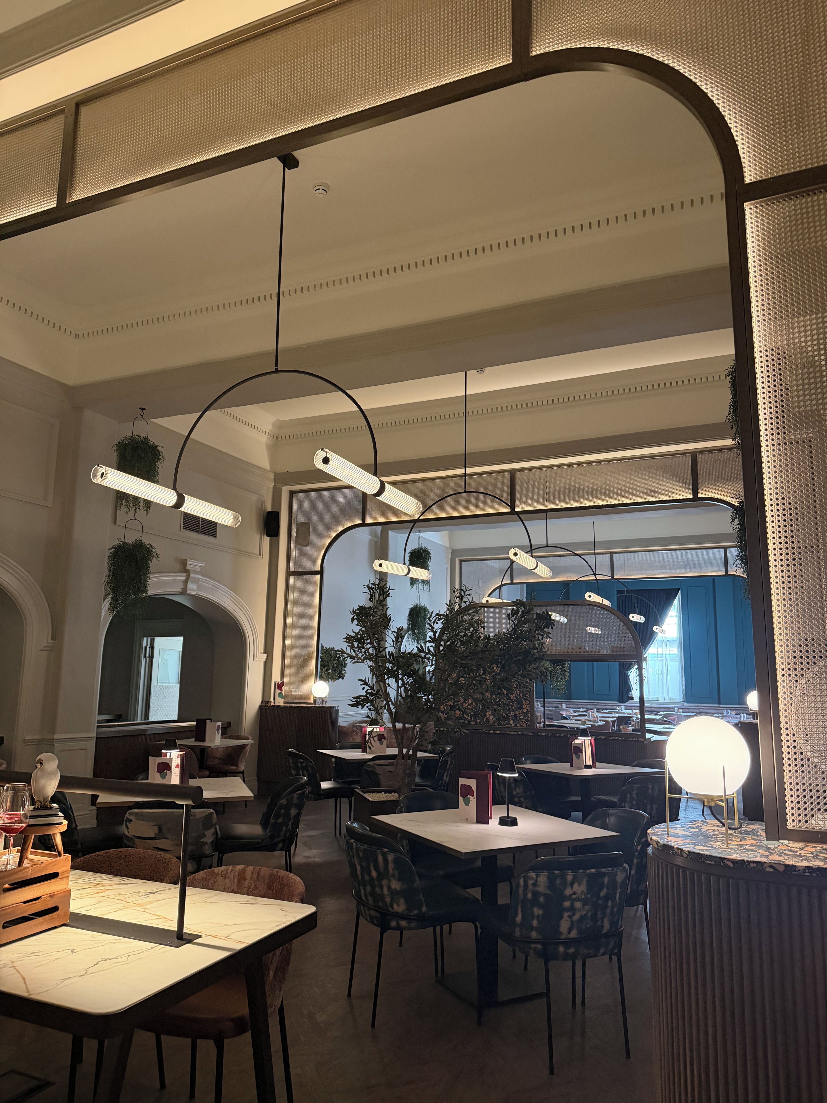 Interior de un restaurante con techos altos, lámparas colgantes y vista de la calle a través de amplios ventanales.