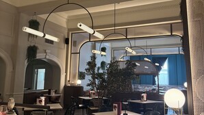 A restaurant interior with high ceilings, hanging pendant lights, and a view of the street through large windows.