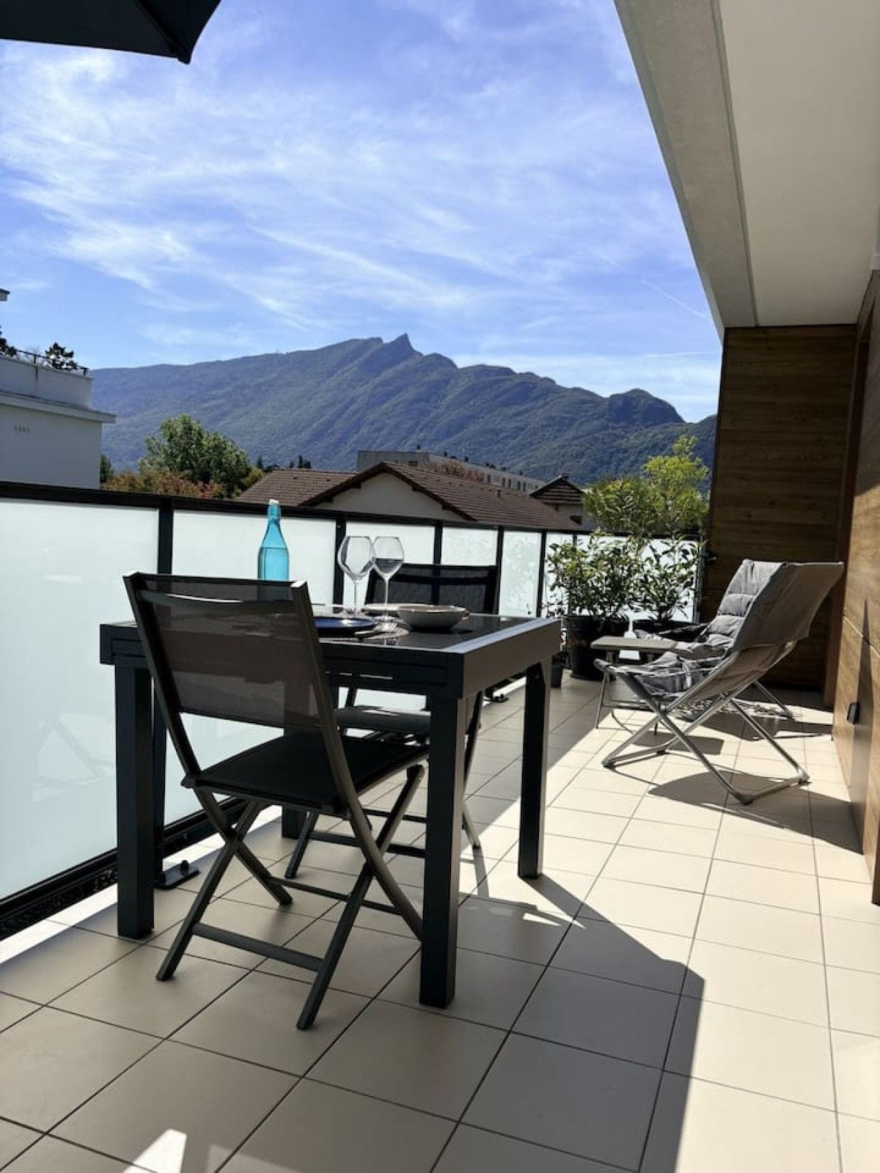 Een balkon met een eettafel en stoelen, een fles en glazen, met uitzicht op een bergachtig landschap.