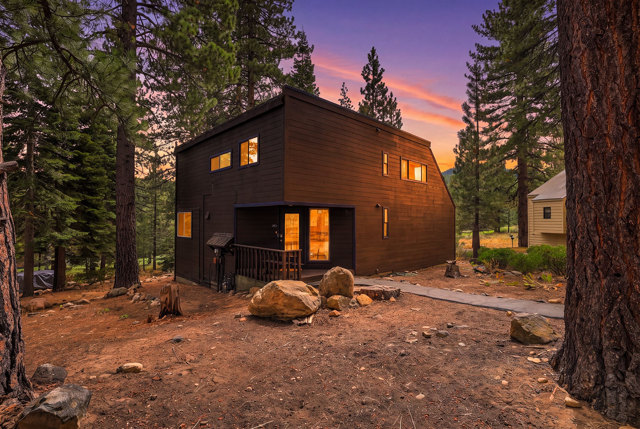A two-story cabin with a dark exterior, surrounded by trees and a dirt ground.