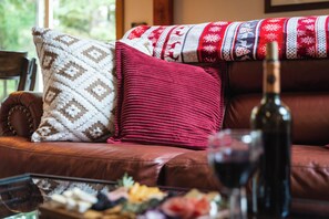 Ein Ledersofa mit gemusterten und einfarbigen roten Kissen, ein Glastisch mit Flasche und Glas, sowie ein Fenster mit Blick auf Grünflächen.