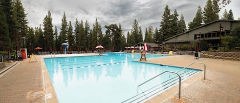 A swimming pool with multiple lanes, surrounded by lounge chairs and umbrellas, with a building in the background.