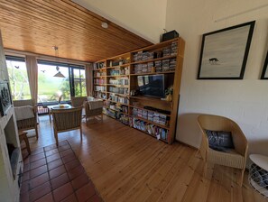 Una zona d'estar àmplia amb sostre de fusta, una gran biblioteca plena de llibres i DVDs, un televisor i mobles de vimet.