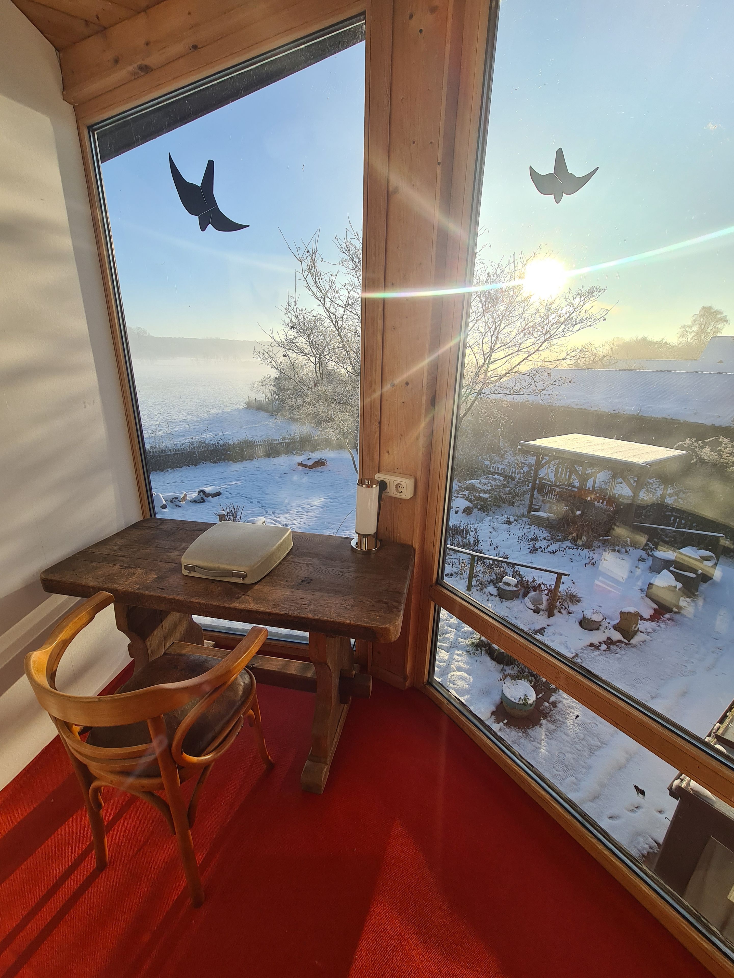Une pièce avec une table et une chaise en bois, un ordinateur portable et une vue sur un paysage enneigé par une grande fenêtre.