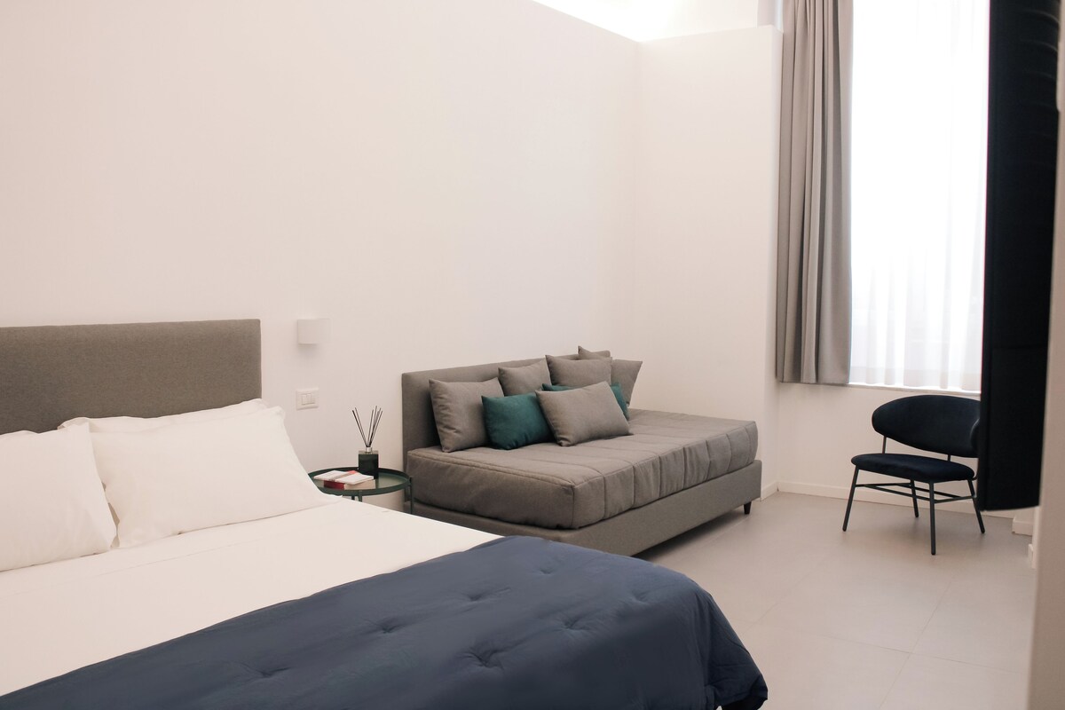 Habitación de hotel moderna con una cama grande, un sofá, una silla y una ventana con cortinas.