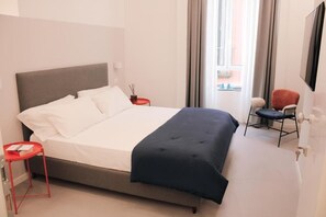 Una habitación de hotel con una cama, una mesita de noche roja, una silla y una ventana con cortinas.