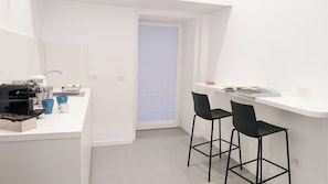Una kitchenette compacta con cafetera, un lavamanos y un ventanal con persianas. Hay dos taburetes negros y una encimera blanca.