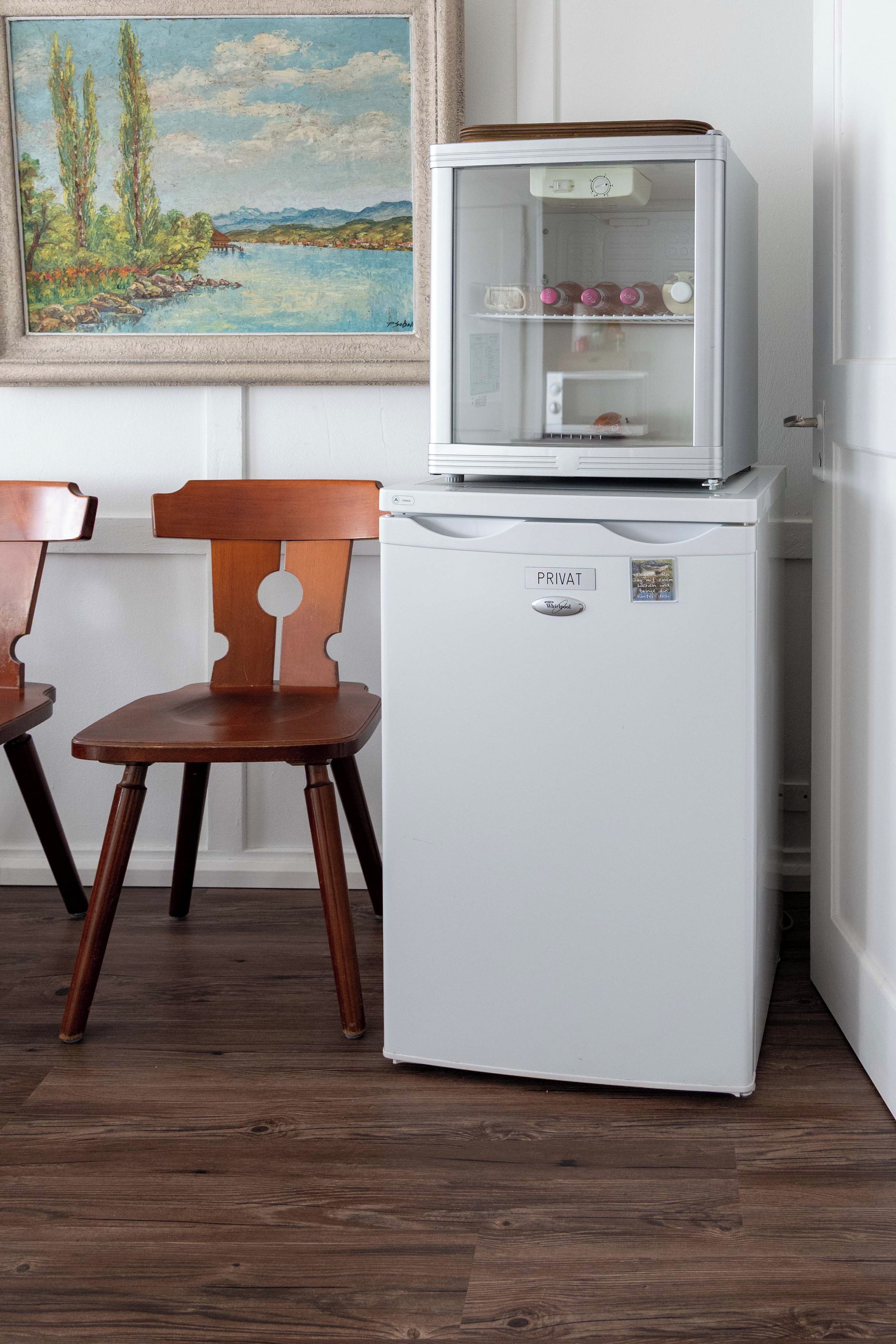 Un refrigerador blanco con puerta de vidrio, una silla de madera y un cuadro en la pared.