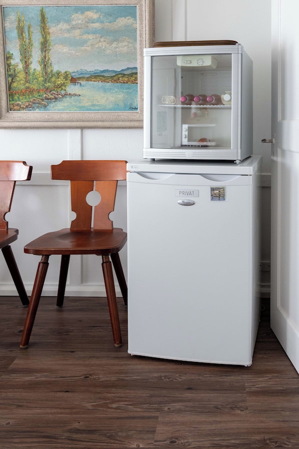 Un refrigerador blanco con puerta de vidrio, una silla de madera y un cuadro en la pared.