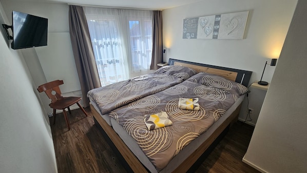 A hotel room with a bed, a chair, a TV mounted on the wall, and a window with curtains.