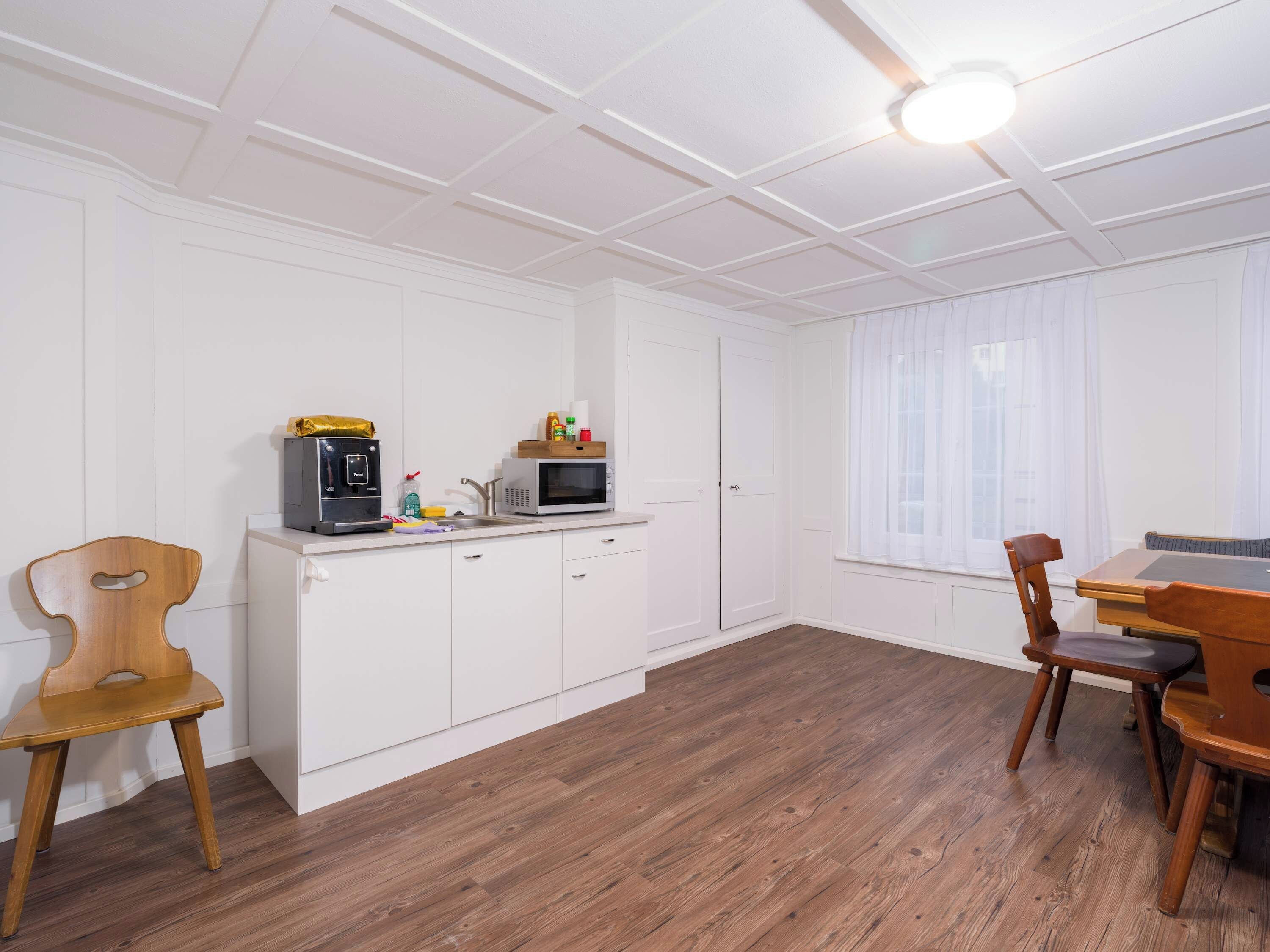 Cocina compacta con muebles blancos, microondas y cafetera. Hay una mesa de comedor con sillas y una ventana con cortinas traslúcidas.