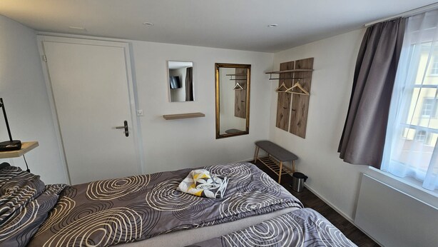 Une chambre à coucher avec un lit, un banc, un miroir et une fenêtre avec des rideaux.