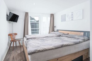 Habitación de hotel con cama, una silla, televisor montado en la pared y una ventana con cortinas.