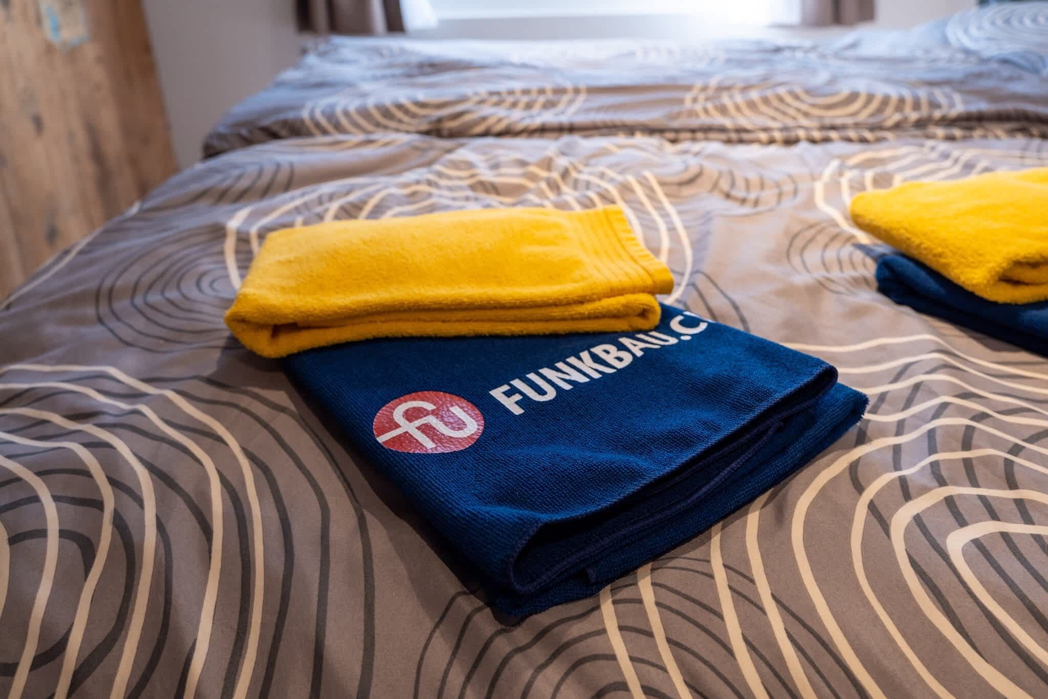 A bed with a patterned bedspread, two folded towels, and a blue towel with a logo.