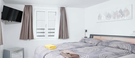 A bedroom with a bed, a television mounted on the wall, a window with curtains, and a small refrigerator.