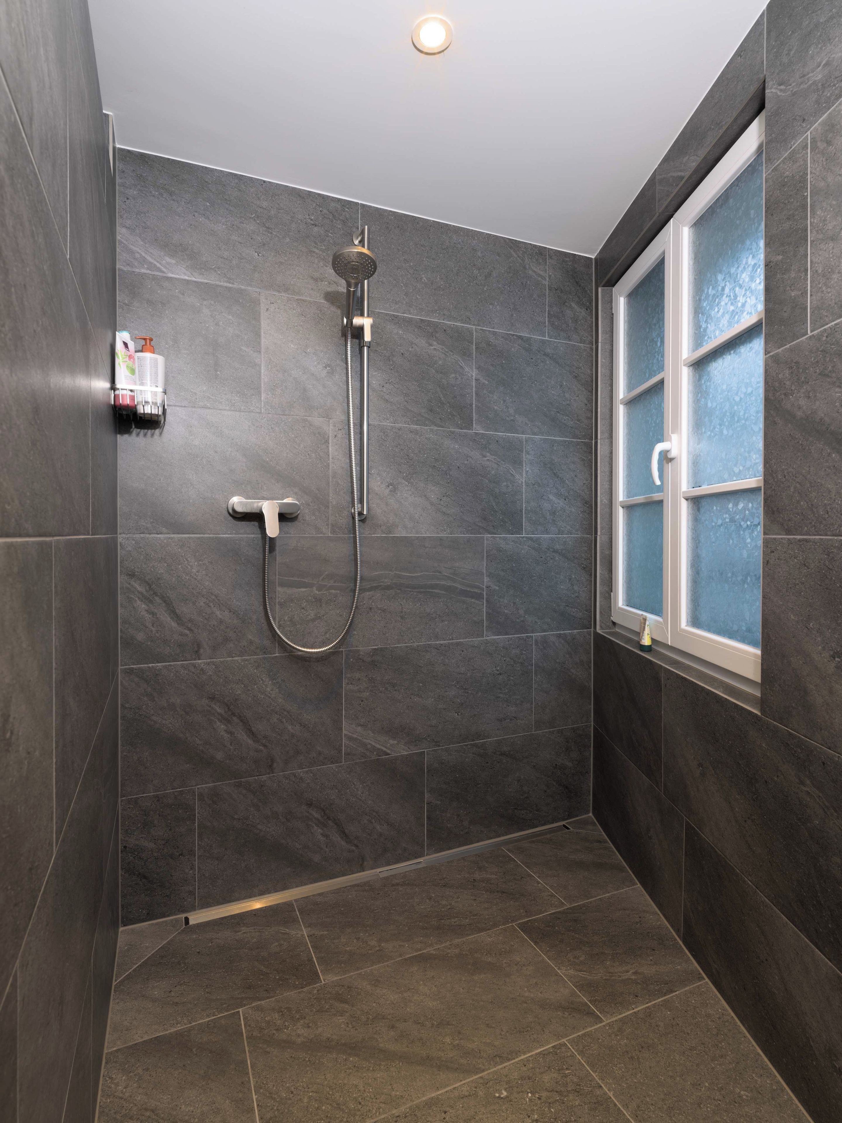 Ein modernes Duschbad mit dunklen Fliesen, einer Handbrause und einem gläsernen Fenster.